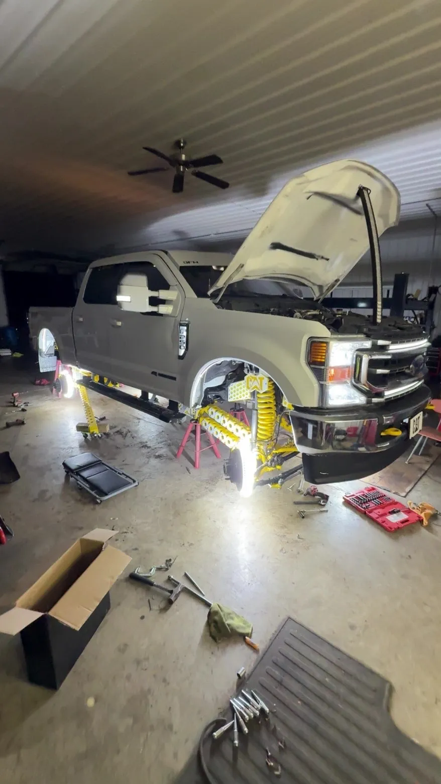 A white Ford F-250 up on jack stands with the hood open, yellow powder-coated suspension components exposed and wheel-well lighting glowing white across the shop floor.
