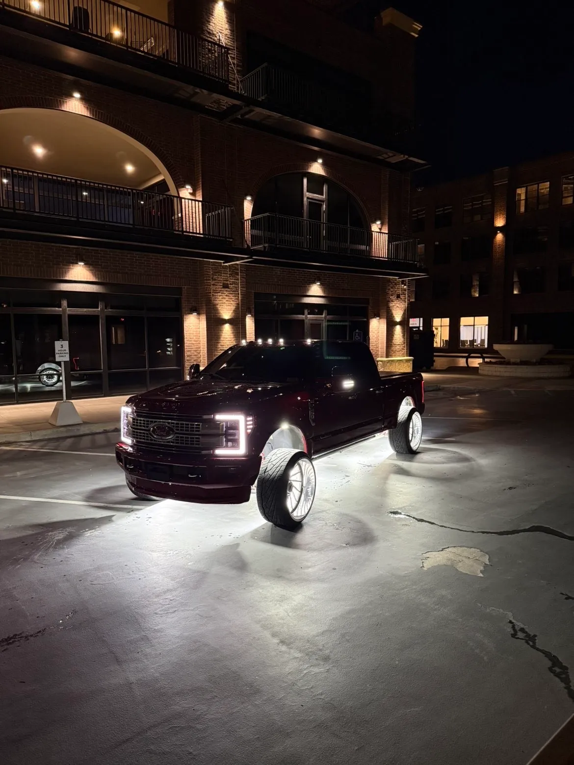 A maroon Ford Super Duty dually parked at night outside a brick building, headlights and LED bars lit, rock lights washing the pavement beneath it.