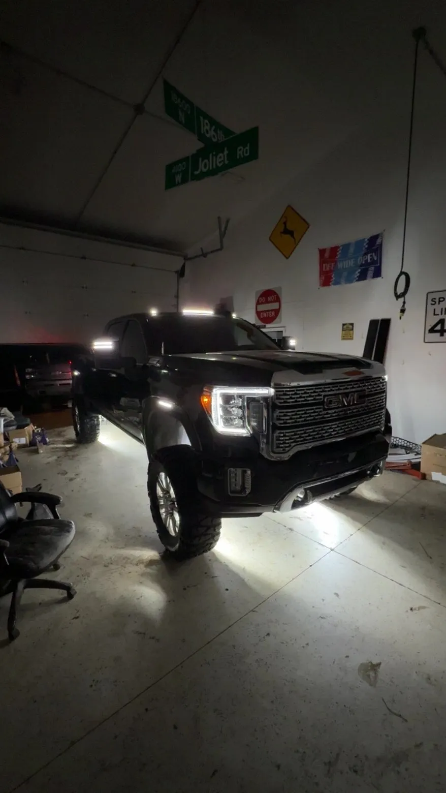 A black GMC Sierra Denali HD lifted on chunky tyres inside the shop at night, rock lights throwing a bright pool of white across the concrete and marker lights lit along the cab roof.