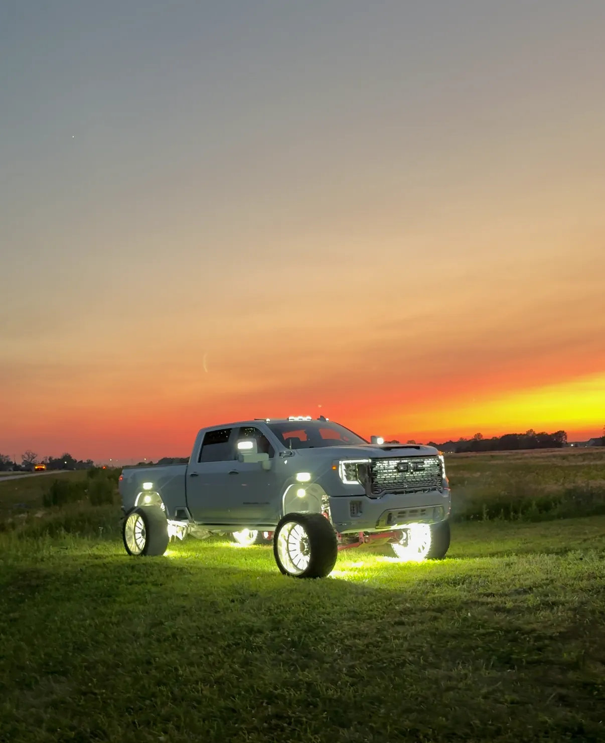 A grey GMC Sierra Denali dually standing in a field at sunset, rock lights blazing white under all four wheels and along the chassis.