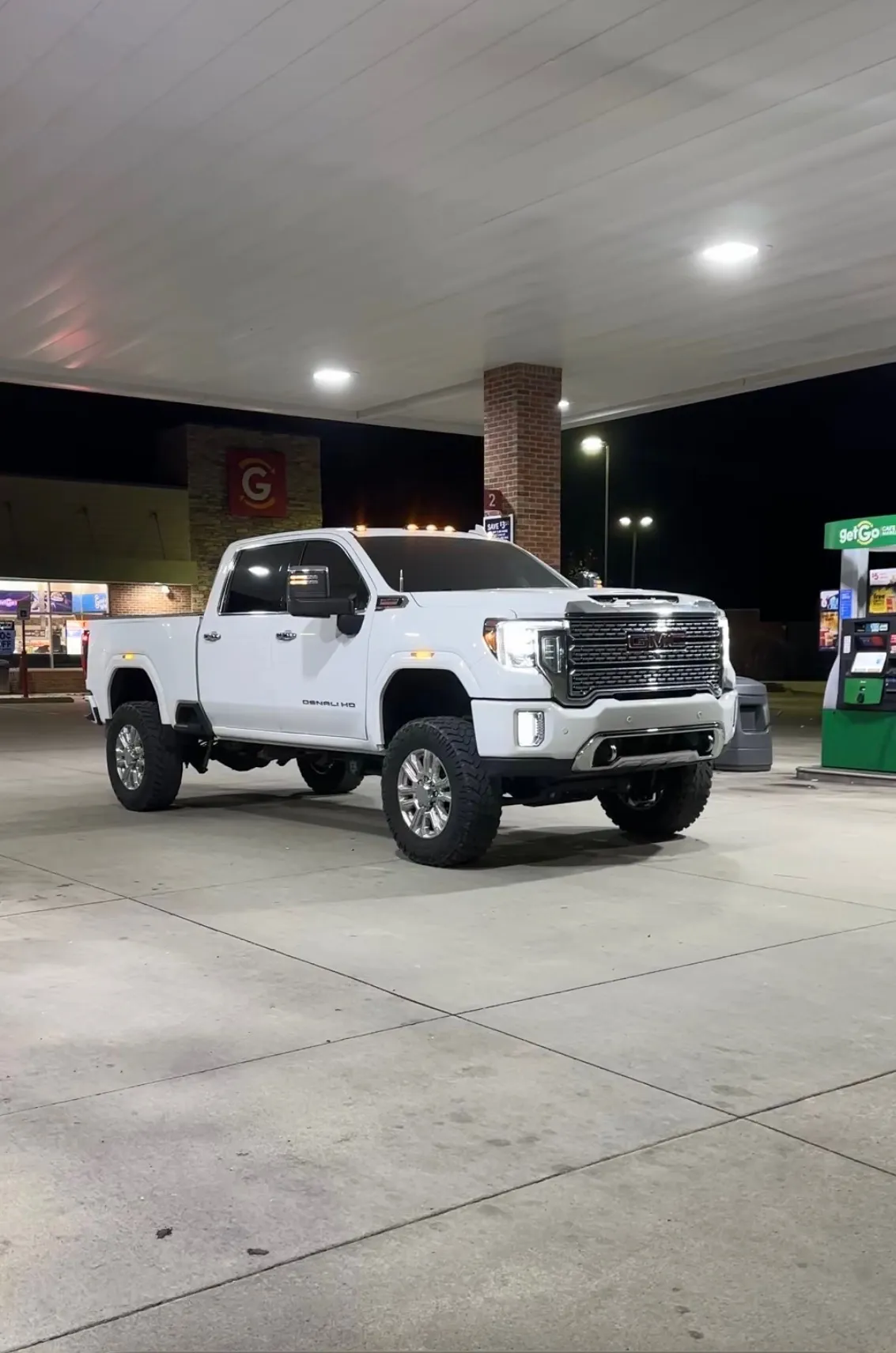 A white GMC Sierra Denali HD under a lit gas station canopy at night, on polished wheels, running only its factory lighting — nothing glowing underneath.